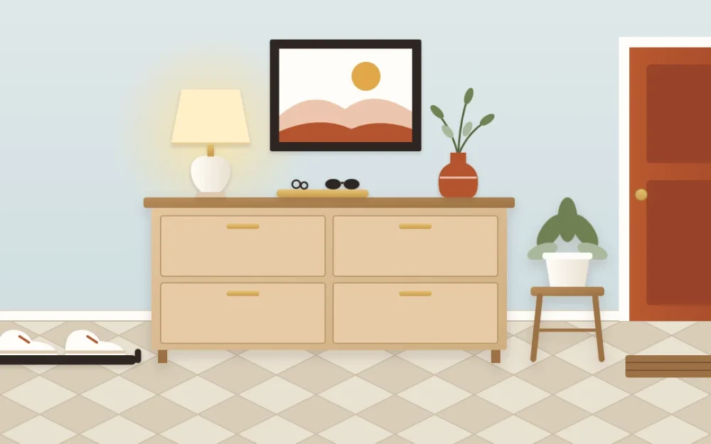 Slim pale wood shoe cabinet styled like a console with a lamp, tray and vase on top, a framed print above and one pair of shoes on a tray