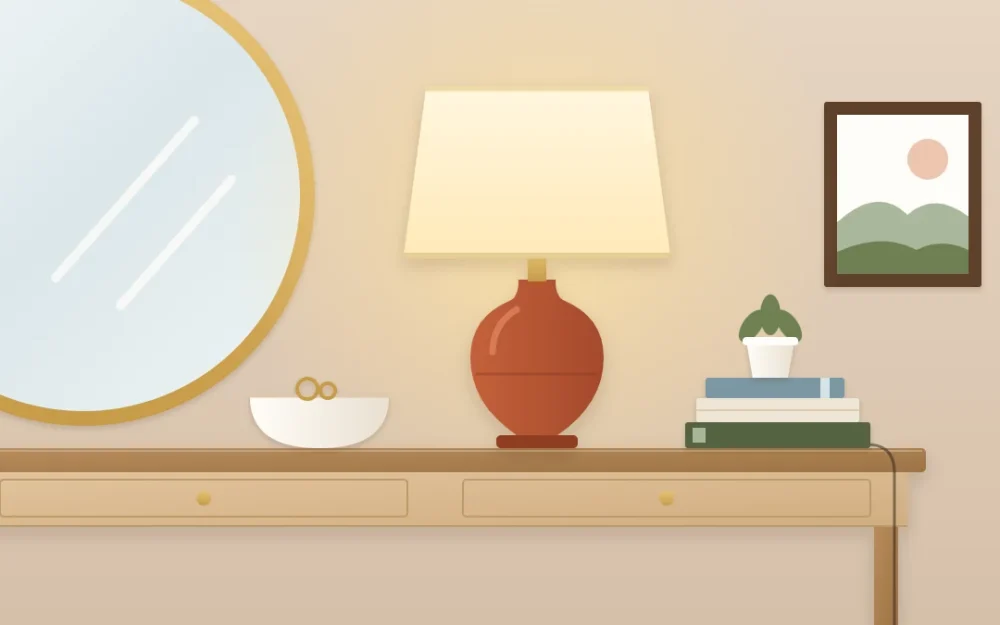One end of a console table with a terracotta ceramic lamp and cream drum shade glowing beside stacked books, a bowl and the edge of a mirror